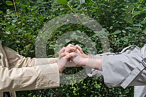 Two pairs of girls hands, dressed in beige and gray jackets, fingers tightly and gently clasped against a background of greenery