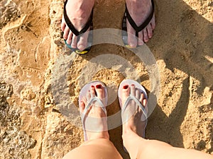 Two pairs of legs male and female with a beautiful pedicure in black and white flip-flops on the sea in a tropical resort on sand