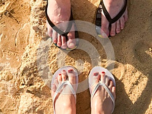 Two pairs of male and female legs with a manicure in slippers, a foot with fingers in flip-flops on a stone sandy floor, earth, be