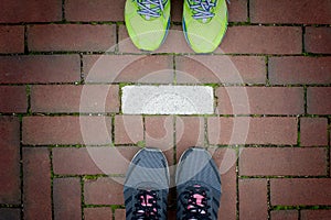 Two pairs of running shoes standing behind each others on red brick walkway with white line separat