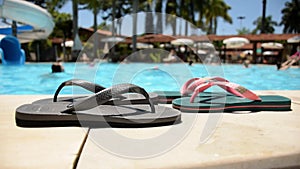 Two pairs of slippers by the pool. In the background people having fun in the water