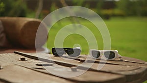 Two pairs of sunglasses, black and white, are lying on a wooden table outdoors