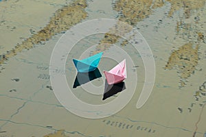 Two paper boats of pink and blue color, float on the water. Under the water you can see a map of the world on which the