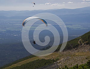 Dvaja paraglajdisti lietajú nad horským údolím za slnečného letného dňa. Kiting v modrom a oranžovom kite v Tatrách