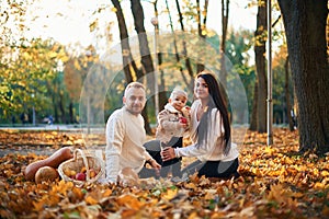 Two parents are sitting with cute little boy in the autumn park on the fallen leaves and butternut squash