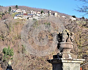 Two pedestals surmounted respectively by heads and eagle in the foreground with a small mountain village in the background.