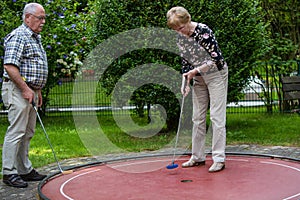 Two pensioners at a minigolf court playing minigolf. He is watch