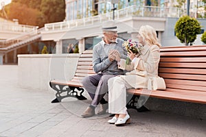 Two pensioners are sitting on a bench in the alley. The aged man gave the woman flowers. He holds her hand