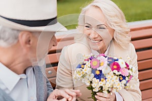 Two pensioners are sitting on a bench in the alley. The aged man gave the woman flowers. He holds her hand