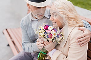 Two pensioners are sitting on a bench in the alley. The aged man gave the woman flowers. He holds her hand