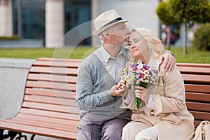 Two pensioners are sitting on a bench in the alley. An elderly man gently kisses a woman on the forehead