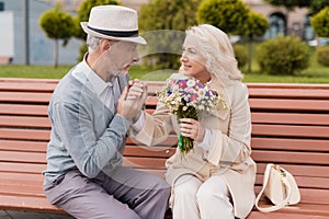 Two pensioners are sitting on a bench in the alley. An elderly man gently kisses a woman`s hand