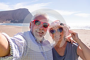 Two pensioners together taking a selfie in the beach smiling and looking at the camera with the sea and the beach at the