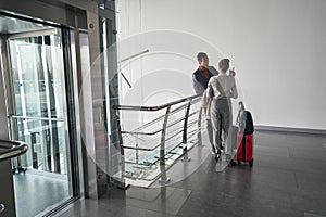 Two people with baggage conversing near lift in corridor