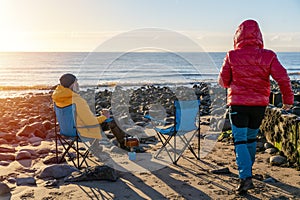 Two people on beach chairs together on winter beach, blue sky horizon. British cold winter. Local tourism concept