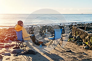 Two people on beach chairs together on winter beach, sunny, blue sky horizon. British cold winter. Local tourism concept