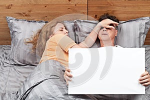 Two people in bed, a woman sleeping with her hand thrown over the man's face, he is holding an empty banner in his hands