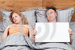Two people are in bed, a woman is sleeping, a man is looking at her, holding a white empty banner facing the camera.