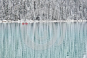 Two people canoeing in red boat on calm blue lake surrounded by winter forest covered with snow.