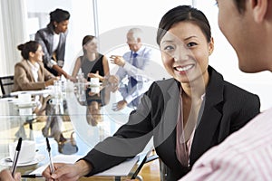 Two People Having Meeting Around Glass Table In Boardroom With Colleagues In Background