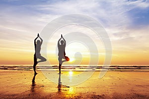 Two people practicing yoga tree position on the beach, sunset