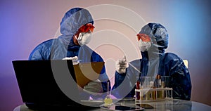 Two people in protective clothing, respirators, glasses and gloves are sitting at a table. they discuss actions, one