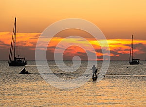 Two people on stand up paddle board at sunset between sailboats