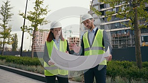 Two people workers builders in safety vests uniform hard hats discuss blueprint document paper plan of unfinished