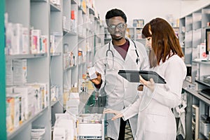 Two pharmacist looking for medicinal drug. Male African pharmacist holding a medicine with female standing by and using