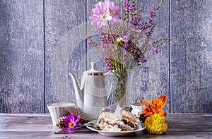 Two pieces of cake in plate, teapot and cups, wild flowers in vase stand on wooden table. Positive mood.
