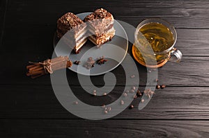 Two pieces of multi-layered honey cake on a plate, on a black wooden background with a cup of tea, chopsticks and coffee