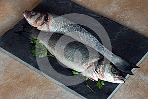Two pieces of sea bass with fresh parsley sprigs on a vintage black slate and a marble table ready to cook.