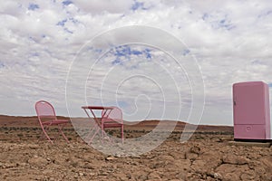 Two pink chairs, a table and a refrigerator stand in the Namib desert against the backdrop of a cloudy sky