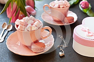 Two pink cups with pink heart shaped marshmallows with macaroons, pink tulips, gift box and silver heart shape pendant on chain.