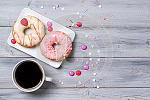 Two pink donuts decorated with smarties and sprinkles hearts and cup of coffee, wooden background