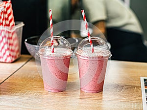 Two pink fruit milk cocktails smoothie in plastic cups with white-red spiral tubes for milkshakes stand on table