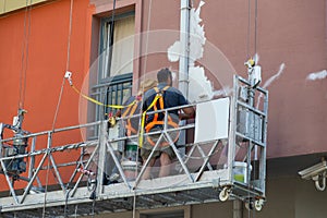Two plasterers painters restoring the facade of the house staying in the building cradle.