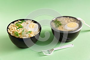 Two plastic bowls of instant noodles decorated eggs and chopped green onion with forks on green background close up