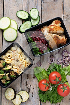 Two plastic  containers with grilled chicken wings and raw vegetables on rustic background, vegetables salad