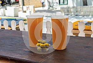 Two plastic half-liter glasses with craft beer and a jar of salted olives stand on a wooden table on the main pedestrian