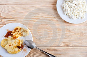 Two plates with curd and cheesecakes with plum jam and metal spoon are standing in opposite corners of wooden table.