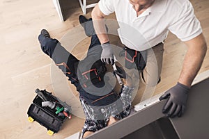 Two plumbers are prepared to repair the water filter. One of them is under the sink, the second is nearby.