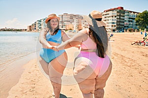 Two plus size overweight sisters twins women happy and proud of their bodies walking at the beach on summer holidays