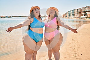 Two plus size overweight sisters twins women happy and proud of their bodies walking at the beach on summer holidays