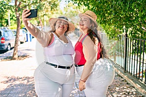 Two plus size overweight sisters twins women smiling taking a selfie picture with the phone outdoors on a sunny day