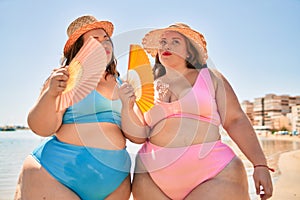 Two plus size overweight sisters twins women using handfan at beach on hoy day of summer