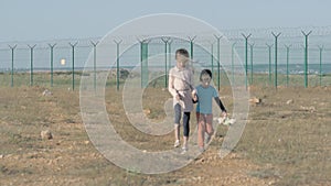 Two poor children refugees family girl and boy with toy walking along desert with razor barbed fence on state border