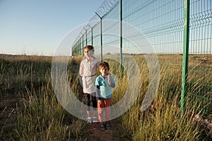 Two poor kids brother and sister refugees standing in green grass near state border with long fence with barbed wire