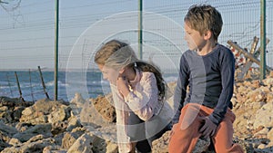 Two poor refugee children boy girl sitting on the stones near the sea. fence barbed wire separating the state.