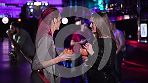 Two positive Caucasian girls standing with cocktails at bar counter, talking and waving to men sitting at the background